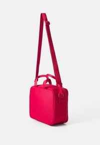 Sac à épaule en tissu rouge de forme rectangulaire, doté de deux poignées courtes et d'une longue sangle ajustable. Texture lisse avec des fermetures éclair.