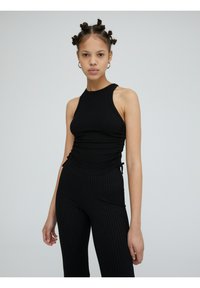 Zwart geribd tanktop met een hoge halslijn en zijruche, gecombineerd met geribde zwarte leggings, waardoor een strakke monochrome outfit ontstaat.
