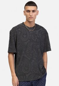 Mørk grå kortærmet t-shirt med struktureret paisley-mønster, rund halsudskæring og løs pasform. Bæres med mønstrede blå bukser.