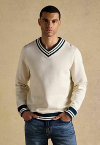 Vyras dėvi baltą megztinį su V formos iškirpte, tamsiai mėlynomis juostelėmis apykaklėje, rankogaliuose ir apačioje, stovintis su rankomis mėlynas džinsų kišenėse priešais smėlio spalvos foną.