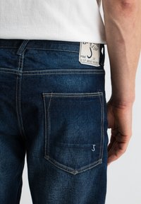 Donkerblauwe denim jeans met een achterzak voorzien van een geborduurd 'J' logo. De tailleband heeft een contrasterend label met tekstdetails.