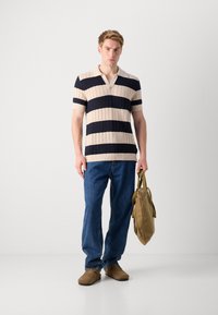 Polo cu mânecă scurtă tricotat, cu dungi bleumarin și bej. Blugi lejeri din denim albastru și pantofi slip-on din piele de căprior maro. Geantă de culoare verde măslinie în mână.