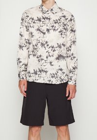 Camicia a maniche lunghe beige con motivi astratti neri, dotata di bottoni sul davanti e colletto, abbinata a pantaloni corti neri fino al ginocchio.