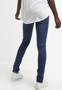 Vaqueros ajustados en denim azul oscuro, con una textura suave, de tiro medio y una silueta ceñida. Combinados con una camisa blanca holgada.