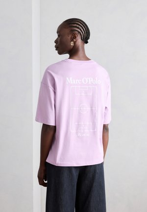 Kvinne med fletter som har på seg en lavendel Marc O'Polo T-skjorte med hvit fotballbane-grafikk og mørke bukser, sett fra siden.