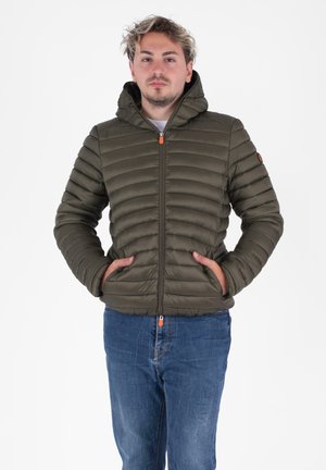 Olijfgroene pufferjack met horizontaal gestikt patroon, voorrits, capuchon en een oranje ritslip, gecombineerd met blauwe jeans.