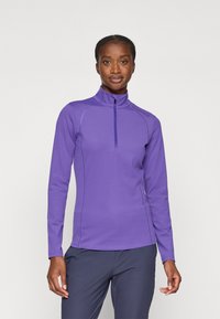 Haut de sport à manches longues violet avec fermeture éclair quart de longueur, coutures à plat, col remonté et tissu lisse et extensible. Porté avec un pantalon sombre.