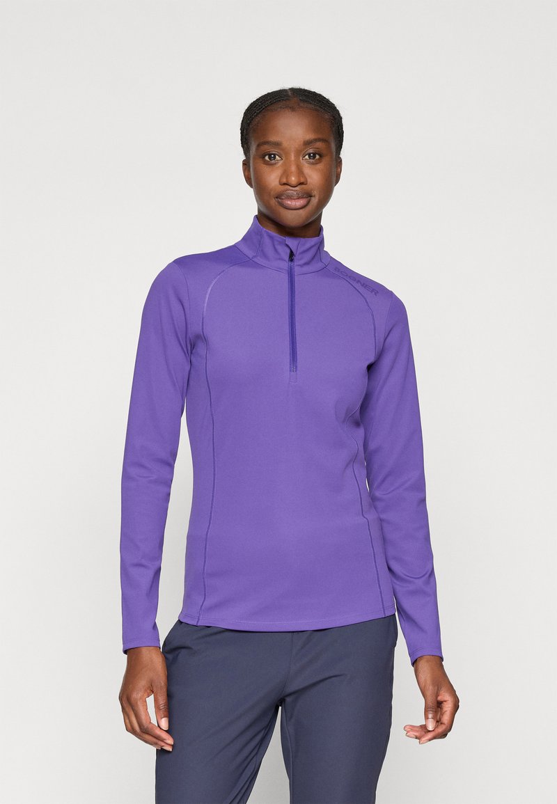 Haut de sport à manches longues violet avec fermeture éclair quart de longueur, coutures à plat, col remonté et tissu lisse et extensible. Porté avec un pantalon sombre.