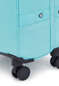 Rolling suitcase in vibrant teal fabric with a textured surface, featuring a black base and wheels for mobility, secured with a webbed strap.