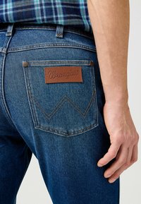 Blauwe denim jeans met een bruine leren logopatch met gestikte details, accenten op de achterzakken en een klassieke snit.