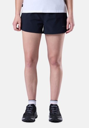 Persona vistiendo pantalones cortos deportivos negros, calcetines blancos hasta el tobillo y zapatillas de correr negras, de pie contra un fondo sencillo.