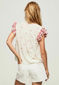 Blouse crème à manches courtes avec des détails floraux rouges brodés et des accents froncés sur les manches, associée à un short en denim blanc.