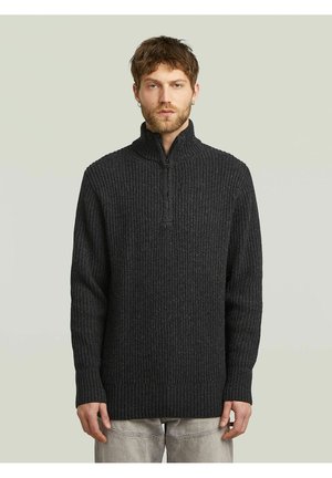 Man met kort bruin haar en baard die een donker geribde gebreide trui met halve ritskraag en lichte broek draagt tegen een effen achtergrond.