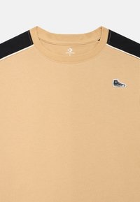 Μπεζ βαμβακερό μπλουζάκι με μαύρα πάνελ στους ώμους, στρογγυλή λαιμόκοψη και μικρό λογότυπο με μαύρο και λευκό αθλητικό παπούτσι στο στήθος.