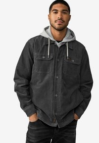 Grå corduroyjakke med to brystlommer, knaplukning og en hættegrå sweatshirt nedenunder. Blød tekstur, afslappet pasform.
