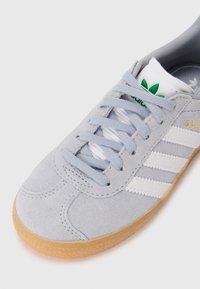 Pelēki zamšādi teniski ar baltām un zeltītām detaļām, ar trim baltām svītrām, apaļu purngalu un gumijas zoli. Redzams zaļš logotipa akcentu.