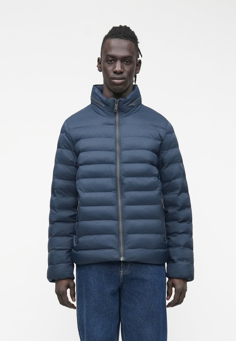 Vīrietis tērpies tumši zilā pūkainā jakā ar priekšējo rāvējslēdzēju un zilās džinsās, stāvot pret baltu vienkrāsainu fonu.