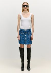 Denim mini-jurk met een knoopsluiting aan de voorkant en twee zakken, gecombineerd met een witte tanktop en hoge zwarte leren laarzen.