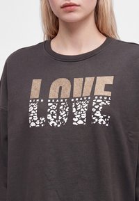 Femme portant un sweat-shirt sombre avec le mot "LOVE" en grandes lettres et un texte plus petit "KIND HEART BRAVE SOUL" au-dessus.