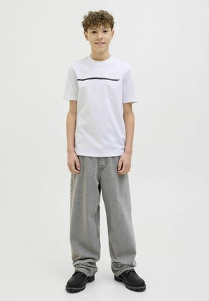 T-shirt en coton blanc avec un motif de ligne noire horizontale, associé à un pantalon en denim gris ample et des chaussures robustes noires.