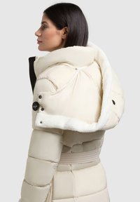 Puffy crèmekleurige jas met grote, donzige capuchon, rits sluiting en knoopaccenten. Heeft een ceintuur in de taille en een geborduurde textuur.