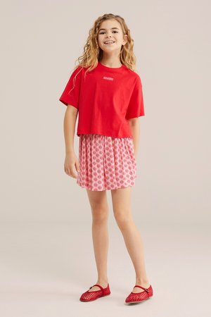 Mosolygós lány hosszú, göndör hajjal, piros rövid felsőt, rózsaszín virágos szoknyát és piros Mary Jane cipőt viselve, világos háttér előtt áll.
