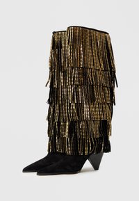 Crne suede čizme do koljena s zlatnim resicama i šiljastim prstima; imaju konusni potpeticu i dekorativne metalne akcentne.