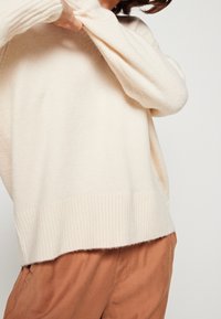 Maglione color crema con maniche lunghe e ampie e orlo a coste. Tessuto morbido, vestibilità rilassata, indossato sopra pantaloni color ruggine.