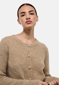 Cardigan beige en tricot avec un col rond, six grands boutons et des poignets côtelés avec une finition douce et texturée.