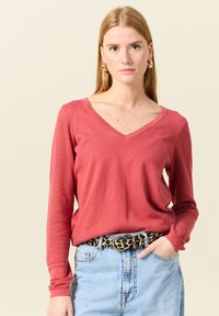 Haut rouge à manches longues avec décolleté en V, présentant des manches en maille texturée et des poignets côtelés. Associé à une jupe en denim taille haute et une ceinture à motif léopard.