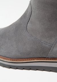 Gros plan sur une botte en daim gris avec des détails cousus et une semelle en caoutchouc superposée avec des accents marron et noir.