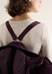 Giacca viola tenuta sopra un maglione beige; presenta spalline regolabili e dettagli con zip, mostrando una texture liscia e leggera.