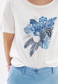 Weißes Baumwoll-T-Shirt mit einem blauen Blumen- und Blätterdruck. Kombiniert mit hellblauen Hosen, die eine glatte Textur und ein einfaches Knopfdetail aufweisen.