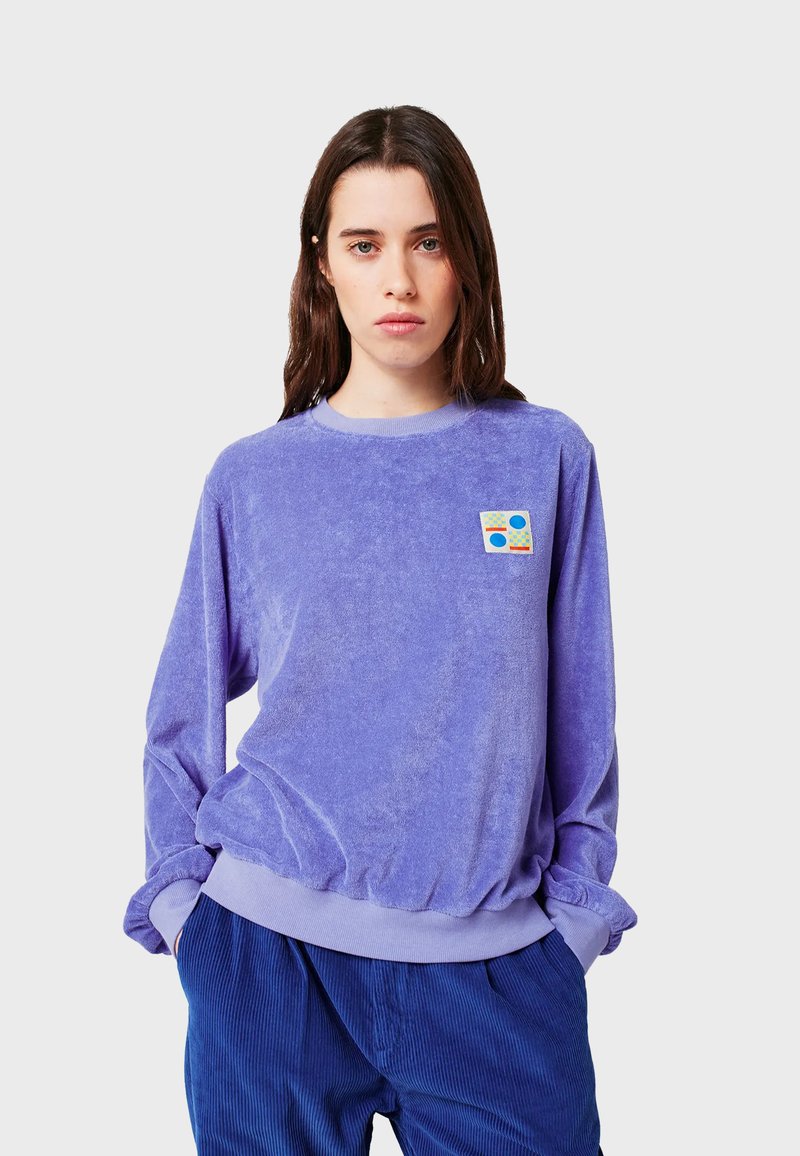 Jonge vrouw met steil bruin haar, gekleed in een lavendelkleurig langmouwen crewneck-top en blauwe corduroybroek, handen in de zakken, neutrale achtergrond.