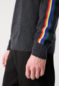 Henkilö, jolla on päällään tummanharmaa villapaita, jossa on sateenkaaren väreissä raitoja hihassa, ja mustat housut. Käsivarsi on rentona sivulla.