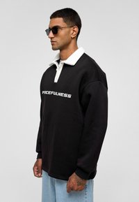 Zwarte sweatshirt met witte kraag en de tekst "VREDIGHEID" in vetgedrukt. Casual fit, katoenmateriaal, lange mouwen en lichte spijkerbroek.