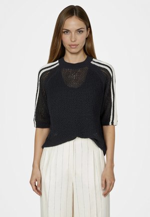 Frau trägt ein schwarzes, kurzärmeliges Strickoberteil mit weißen Streifen auf den Schultern und weiß gestreifte, hoch taillierte Hosen vor schlichtem Hintergrund.