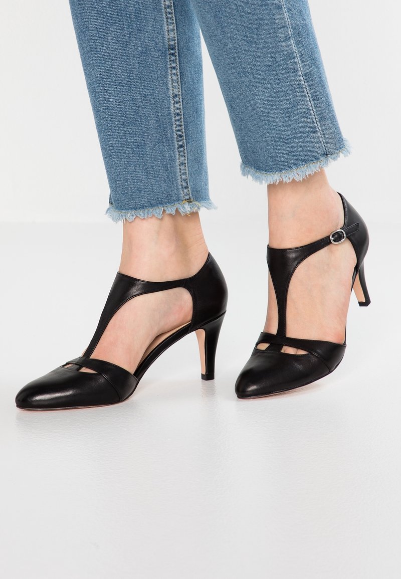 Escarpins à talons hauts noirs en cuir avec bride en T, portés avec un jean court bleu clair à ourlet effiloché sur un sol blanc.