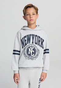 Felpa grigia con stampa blu marina, che presenta "NEW YORK" e un emblema universitario. Maniche lunghe con strisce blu marina. Abbinata a pantaloni della tuta grigi.
