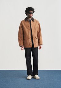 Καφέ μπουφάν με κουμπιά και σκούρο γιακά, δύο μπροστινές τσέπες, φορεμένο πάνω από μαύρο πουκάμισο και μαύρο παντελόνι, συνδυασμένο με ανοιχτόχρωμα αθλητικά παπούτσια και γυαλιά ηλίου.