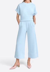 Ljusblå tvådelad set med en avspänd topp med uppvikta ärmar och vida byxor med sidofickor och detaljer vid fållen. Rosa spetsiga skor.