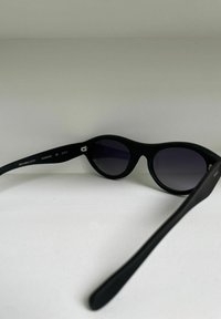 Occhiali da sole neri con forma arrotondata, lenti scure e finitura opaca. Caratteristiche includono un piccolo logo sulla tempia e una classificazione UV di Categoria 3.
