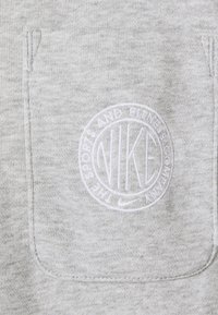 Grått tyg med en texturerad yta, med en broderad vit Nike-logotyp i en cirkulär design, omgiven av orden "THE SPORT AND FITNESS COMPANY."