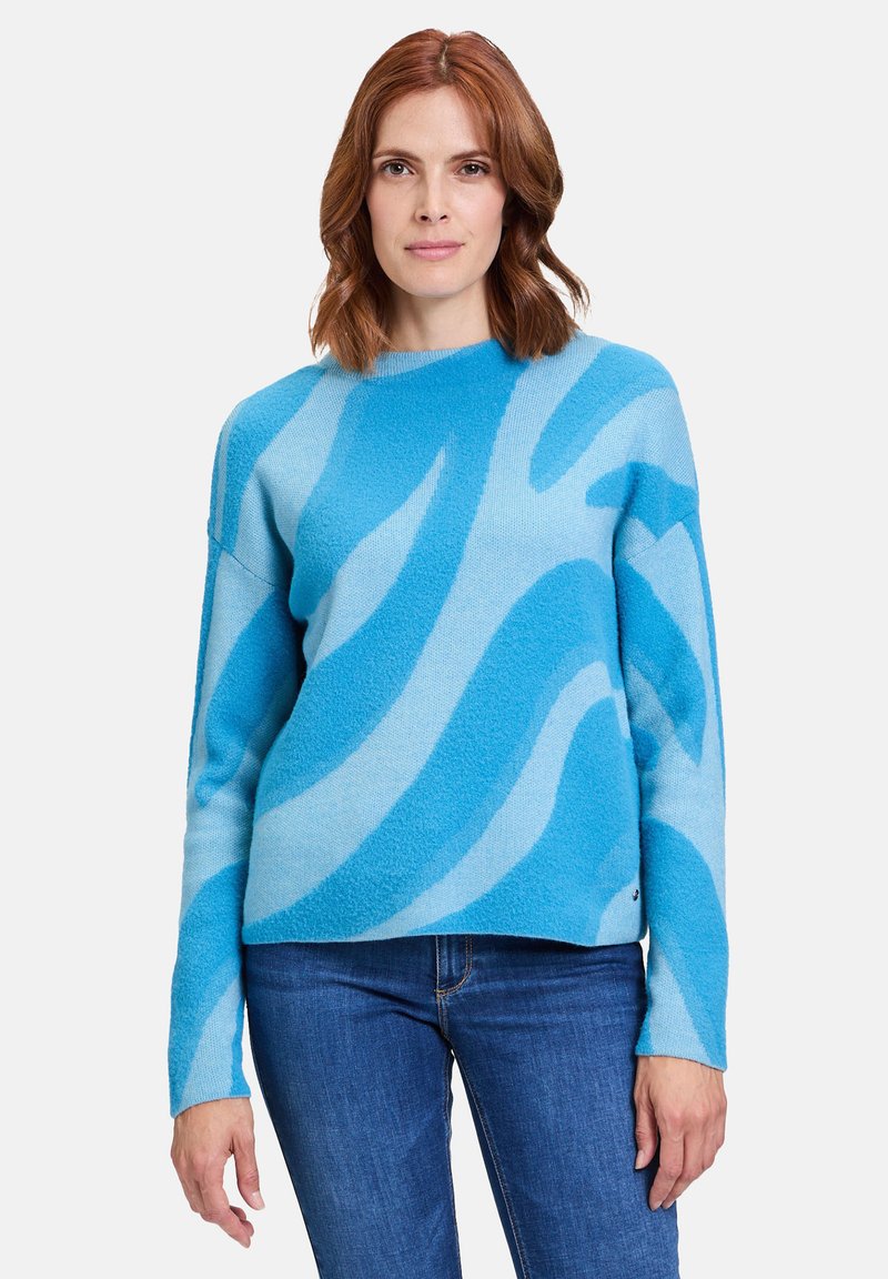 Blauer Fleece-Pullover mit hellblauen Wellenmustern, Rundhalsausschnitt und langen Ärmeln; kombiniert mit Jeans.