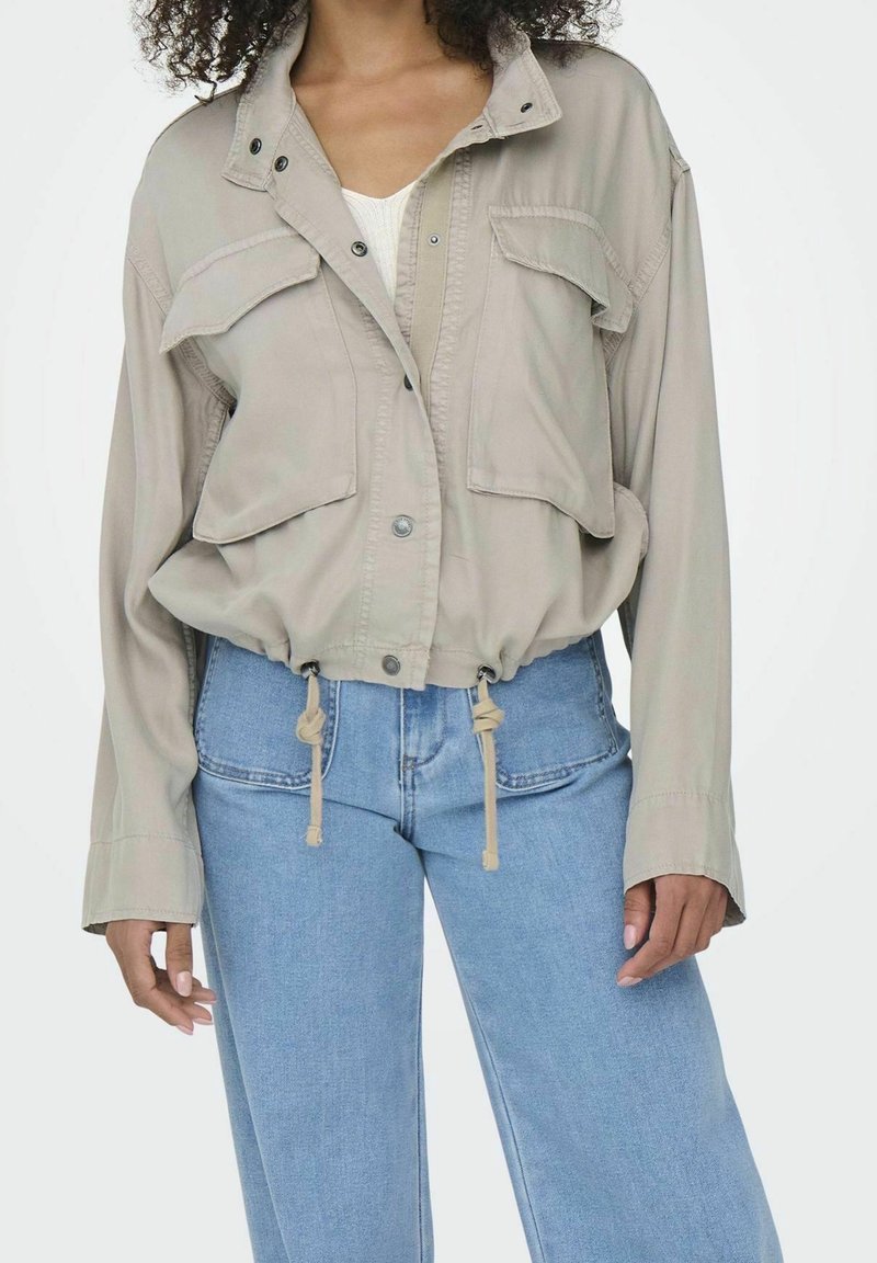 Beige arbetsjacka med två bröstfickor, tryckknappar, dragsko i midjan och långa ärmar, tillsammans med ljusblå vida jeans.