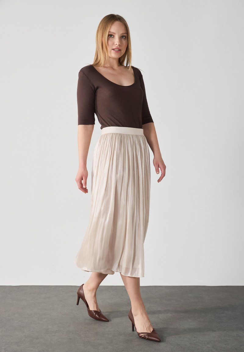 Top marrón ajustado de manga larga combinado con una falda beige plisada y fluida, y zapatos de tacón marrones con punta, resaltados contra un fondo neutro.