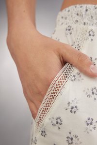 Floral patroon stof in crème kleur met kantafwerking bij de zak. De hand is zichtbaar in de zak gestoken, wat het ontwerp detail benadrukt.