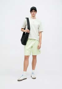 Cămașă scurtă gri deschis cu logo, pantaloni scurți verzi neon, pantofi albi cu accente gri, șapcă neagră și o geantă neagră de umăr.