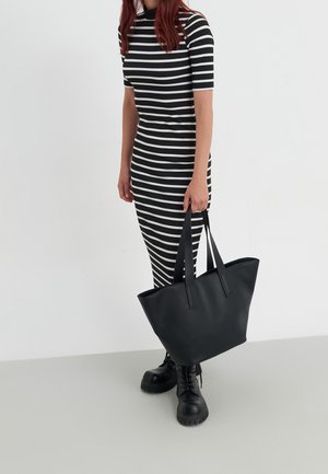 Femme portant une robe à rayures noires et blanches, des bottes noires épaisses, et tenant un grand sac fourre-tout en cuir noir sur un fond blanc uni.