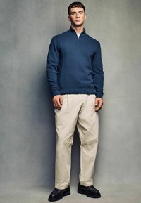 Maglione a collo alto a zip quarti di colore blu navy, pantaloni beige con pieghe e scarpe nere con suola spessa. Texture liscia, vestibilità su misura e minimalismo nei dettagli.